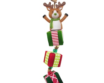 May include: A hanging ornament featuring a brown reindeer with a green scarf, positioned above three stacked cube-shaped gift boxes. The top box is green with red stripes, the middle is red with white dots, and the bottom is dark green with a pink pattern.