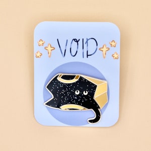 May include: A black and gold enamel pin featuring a cartoon cat with a starry body and a gold box on its head. The pin is on a blue card with the word "VOID" written in black and white stars on either side.