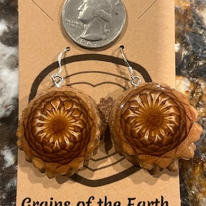 May include: A pair of earrings with a unique design. The earrings are made from a natural material, possibly a pinecone, with a detailed, circular pattern. The earrings are attached to silver-colored hooks. The card behind the earrings has the text "Grains of the Earth".