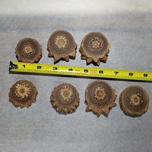 May include: Seven brown, circular, wooden pine cone cross sections with a detailed, intricate, carved pattern. The pine cone cross sections are arranged in two rows on a white surface with a yellow measuring tape.