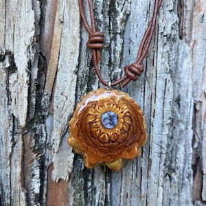 May include: A handcrafted pendant necklace featuring a natural, flower-shaped pendant with a central blue stone. The pendant is suspended from a brown leather cord, knotted at the top. The pendant is approximately 1.5 inches in diameter.