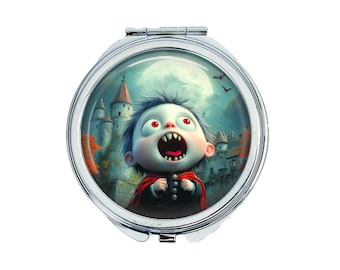 May include: A silver compact mirror with a round image of a cartoon vampire baby with red eyes and fangs, wearing a red cape and black clothing, against a backdrop of a castle and a full moon.
