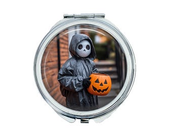 May include: A silver compact mirror with a round image of a child dressed in a gray hooded costume holding an orange pumpkin. The child is wearing a white skull mask with black eyes.