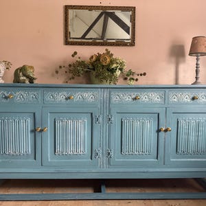 May include: A teal-painted sideboard with four doors and ornate detailing. The cabinet features gold-toned handles and stands on a solid base. A mirror and decorative items, including a lamp, are displayed on top.