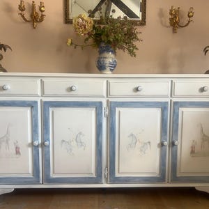May include: A white sideboard with four doors and four drawers. The doors are framed in blue and feature hand-painted illustrations of giraffes and horses. The sideboard is topped with a vase of flowers and decorative candle holders.