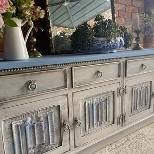 May include: A large, grey and blue painted sideboard with multiple drawers and cabinet doors. The cabinet doors have a carved design. The top of the sideboard is blue, and the edges are trimmed with gold. The sideboard is decorated with flowers and a mirror.
