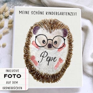 Könnte beinhalten: Ein weißer Ordner mit einer Aquarell-Illustration eines Igels, der eine Brille trägt. Der Text "Meine schöne Kindergartenzeit" befindet sich oben auf dem Ordner. Der Text "Pepe" ist unter dem Igel geschrieben. Der Text "INKLUSIVE FOTO AUF DEM ORDNER-RÜCKEN" befindet sich in einem weißen Kasten mit schwarzem Rand.