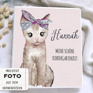 Könnte beinhalten: Ein personalisierter Ordner mit einer Aquarellillustration einer Katze mit Schleife. Der Umschlag ist hellrosa mit dem Namen "Hannah" und deutschem Text. Inklusive Foto auf dem Rücken.