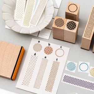 May include: A collection of wooden stamps with various geometric patterns including stripes, dots, and grids. A white paper sheet displays stamped impressions labelled a through f, alongside loose paper strips and square samples with matching designs.