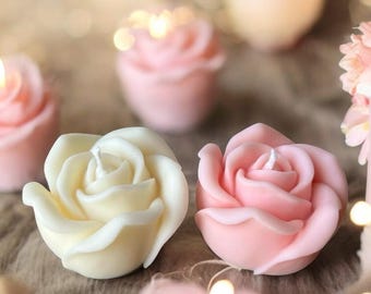 Könnte beinhalten: Nahaufnahme von rosenförmigen Kerzen in zarten Rosa- und Cremefarben. Die Kerzen haben ein realistisches Blütenblattdesign und einen weißen Docht. Der Hintergrund ist mit Bokeh-Lichtern verschwommen, was eine warme und romantische Atmosphäre schafft.