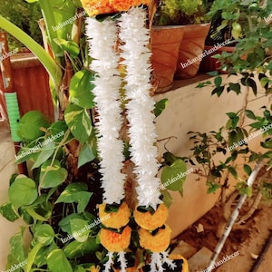 May include: Two white flower garlands with yellow flower accents. The garlands are hanging from a tree branch. The garlands are made of artificial flowers.