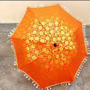 May include: An orange and gold umbrella with a floral pattern and tassels. The umbrella is made of fabric and has a wooden handle.