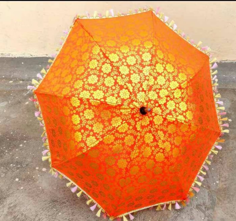 May include: An orange and gold umbrella with a floral pattern and tassels. The umbrella is made of fabric and has a wooden handle.
