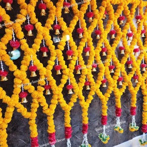 May include: A decorative wall hanging with bright yellow marigold garlands in a diamond pattern. Red and white accents, small golden bells, and green and yellow floral clusters add detail. The text "indiangarlanddecor" is visible.