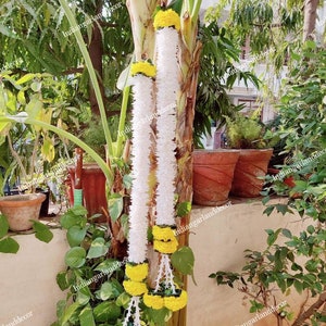 May include: Two white flower garlands with yellow marigold accents hanging on a tree trunk. The garlands are made of artificial flowers and are used for decoration.