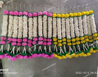 May include: A collection of white flower garlands with pink and yellow pom-poms at the ends. The garlands are hanging from a string and are arranged in rows. The text "Indiangarlanddecor" is visible on the image.