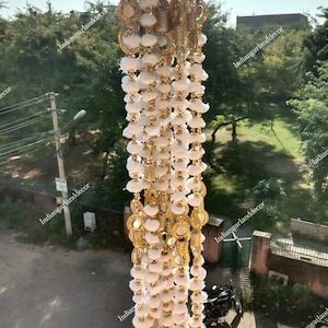 Puede incluir: Guirnalda de cuentas blancas y doradas con pequeños espejos redondos. La guirnalda está colgada de una ventana y tiene un borla en la parte inferior.