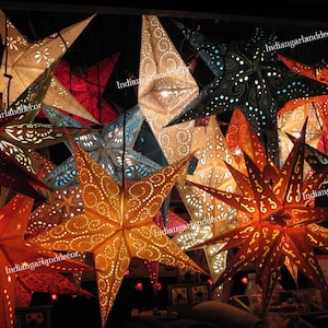 Op de afbeelding: Een verzameling kleurrijke papieren stervormige lantaarns met ingewikkelde ontwerpen. De lantaarns worden van binnenuit verlicht, waardoor een warme en feestelijke sfeer ontstaat. De tekst "indiangarlanddecor" verschijnt meerdere keren op de afbeelding.