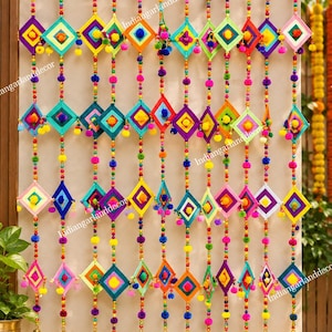 Op de afbeelding: Een wandhanger met meerdere verticale strengen van kleurrijke kralen, kleine pompons en ruitvormige stoffen ornamenten in diverse felle kleuren. Elke streng eindigt aan de onderkant met een kleine goudkleurige metalen bel.