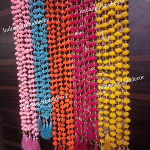 Puede incluir: Una colección de guirnaldas coloridas, cada una con un tono único. Las guirnaldas presentan pompones y borlas en tonos rosa, azul, naranja, fucsia y amarillo. El texto "indiangarlanddecor" es visible en la imagen.