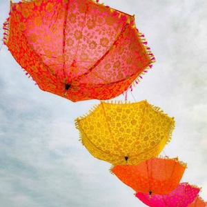 May include: Four colorful umbrellas with floral patterns hang from the ceiling against a cloudy sky. The umbrellas are pink, yellow, orange, and pink.