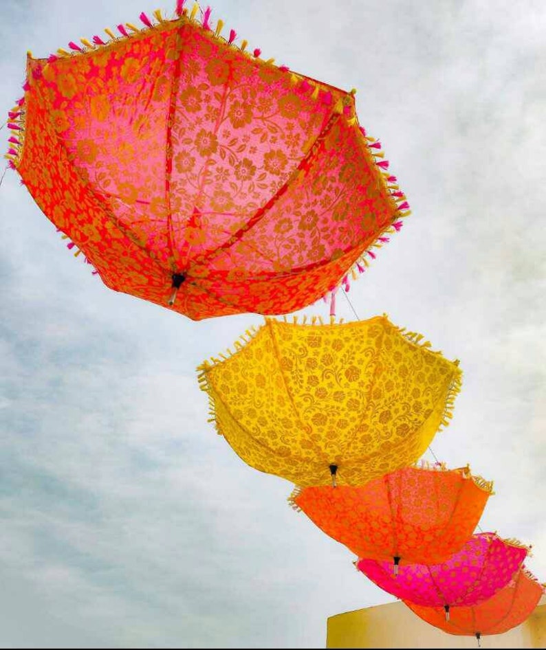 May include: Four colorful umbrellas with floral patterns hang from the ceiling against a cloudy sky. The umbrellas are pink, yellow, orange, and pink.