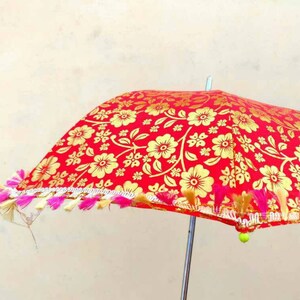 May include: A red umbrella with a gold floral pattern and pink and white fringe trim. The umbrella has a silver handle and a green tassel.