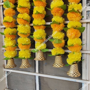 Puede incluir: Cinco guirnaldas de flores amarillas y naranjas con campanas doradas colgando de una ventana. Las guirnaldas están hechas de flores de tela y hojas verdes. El texto "IndianGarlandDecor" es visible en la imagen.