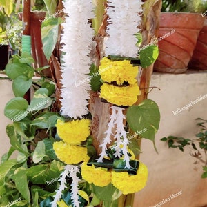 May include: Two white flower garlands with yellow marigold flower garlands hanging on a tree trunk. The garlands are tied together with green leaves. The image has the text "indiangarlanddecor" on it.