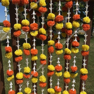 May include: A garland of yellow and orange artificial marigold flowers strung together with white jasmine flowers. The garland is hanging on a wooden fence.