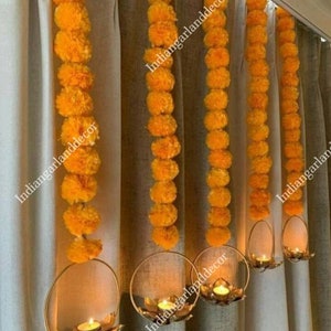 Pode incluir: Cinco guirlandas de flores de calêndula amarelas penduradas em uma cortina com castiçais de metal dourado. Cada castiçal tem uma vela de chá acesa.