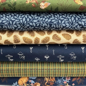 Op de afbeelding: Een stapel verschillende stoffen met patronen. Ontwerpen omvatten bladeren, dennenappels, paddenstoelen en ruitjes in groen, blauw en beige tinten. De stoffen zijn waarschijnlijk van katoen of een vergelijkbaar materiaal, geschikt voor patchwork.