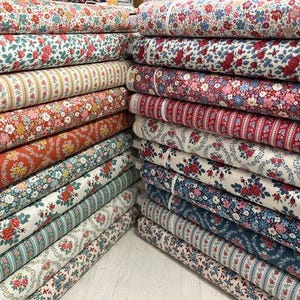 Könnte beinhalten: Eine Sammlung von Stoffballen, die in zwei Säulen gestapelt sind und eine Vielzahl von Blumen- und Streifenmustern in Rot-, Blau-, Grün- und Cremetönen aufweisen. Die Textilien zeigen komplexe botanische Drucke, kleine Rosenmotive und vertikale Streifen.