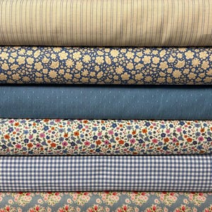 Op de afbeelding: Een stapel verschillende stoffen rollen in tinten blauw, beige en wit. Patronen zijn onder meer strepen, bloemenprints, stippen en ruitjes. De stoffen lijken van katoen of een vergelijkbaar materiaal te zijn, geschikt voor naai- en knutselprojecten.