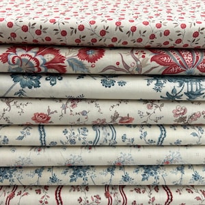 May include: A pile of patterned fabrics in a variety of floral designs. The fabrics display patterns in red, blue, and cream. Some designs include small red flowers, large floral motifs, and striped borders with floral accents. The fabrics appear to be cotton or linen.