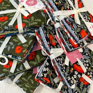 May include: A collection of folded fabric bundles tied with white ribbons. The fabric features botanical prints with red poppies, white flowers, and green foliage on dark olive green and black backgrounds, including tags labelled Fat Quarter Pack.