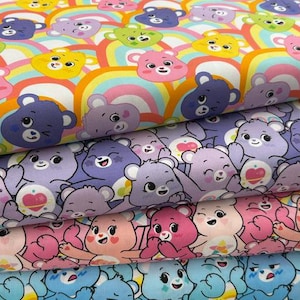 Peut inclure: Une pile de trois rouleaux de tissu pliés représentant des personnages d'ours de dessin animé. La couche supérieure affiche des arcs-en-ciel colorés et des visages d'ours en rose, jaune, vert et violet. La couche intermédiaire montre des ours violets avec des symboles de cœur. La couche inférieure présente des ours roses et bleus.