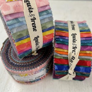 May include: A collection of fabric bundles in vibrant colours and patterns. The image shows a rolled bundle and two rectangular stacks, each bound with a cream ribbon printed with "Lewis & Irene." The fabrics display a range of hues.
