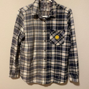 May include: A long-sleeved flannel shirt with a split design. One half is gray and white plaid, the other is navy and white plaid. A yellow smiley face patch is on the chest pocket. The shirt is hanging on a white hanger.