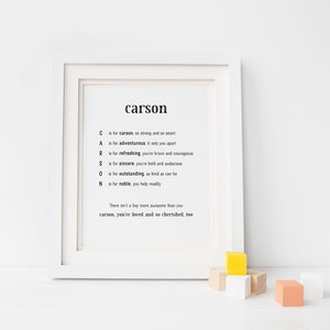 Puede incluir: Un marco blanco con una impresión del nombre "carson" en negro, acompañado de cualidades positivas. Pequeños bloques de madera de colores están frente al marco. El texto describe las cualidades del nombre.
