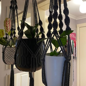 May include: Three hanging plant macrame planters with black, white, and grey pots. The macrame is made of grey rope and has a tassel at the bottom. The plants are green and leafy.