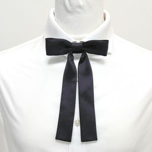 May include: A black satin bow tie with long, flowing ends, set against a white dress shirt. The bow tie is centered and secured with a small button. The overall look is formal and elegant.