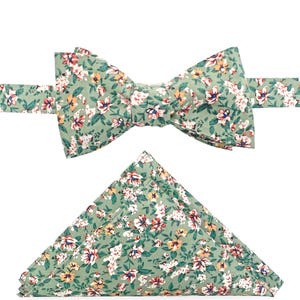 Op de afbeelding: Een groene vlinderdas en bijpassende pochet set met bloemenmotief. De vlinderdas is voorgeknoopt en heeft een patroon van kleine witte, gele en rode bloemen op een groene achtergrond. De pochet is in een driehoek gevouwen.