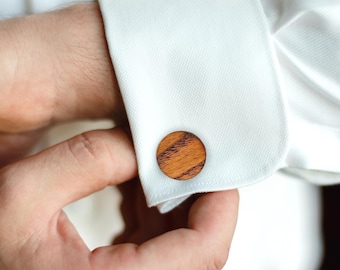 May include: A pair of round wooden cufflinks with a brown and reddish-brown wood grain pattern.