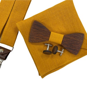Puede incluir: Un conjunto de tirantes de color amarillo mostaza, una pajarita de madera y gemelos. La pajarita está hecha de madera oscura y tiene una banda de tela amarilla mostaza. Los gemelos son de plata.