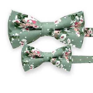 Op de afbeelding: Twee groene vlinderdassen met een bloemenpatroon van witte en roze bloemen. De vlinderdassen zijn gemaakt van stof en hebben een klassieke strikvorm. De achtergrond is wit.