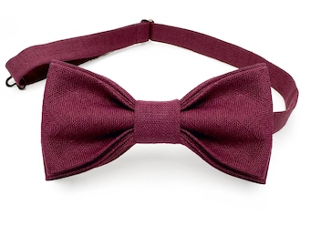 May include: A burgundy bow tie made of a textured fabric. The bow tie is attached to a burgundy adjustable strap.