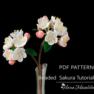 May include: A PDF pattern for a beaded sakura flower tutorial. The image shows a beaded flower arrangement in a clear glass vase against a black background. The flowers are white with pink accents and yellow centers. The text "PDF PATTERN Beaded Sakura Tutorial Anna Masalska" is visible at the bottom of the image.
