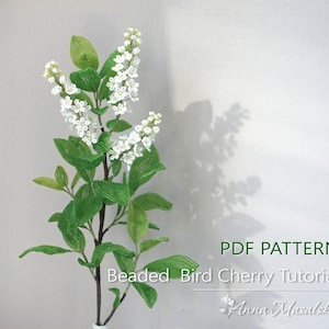 Può includere: Un tutorial di Bird Cherry con perline. L'immagine mostra un gambo floreale dettagliato con perline con fiori bianchi e foglie verdi. Il testo "PDF PATTERN" e "Beaded Bird Cherry Tutorial" sono visibili. Viene visualizzato anche il nome dell'artista, Anna Masalska.
