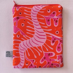Può includere: Una pochette con cerniera rossa e rosa con un motivo astratto fantasioso. La pochette ha il testo "while sadie sleeps" stampato sul lato.